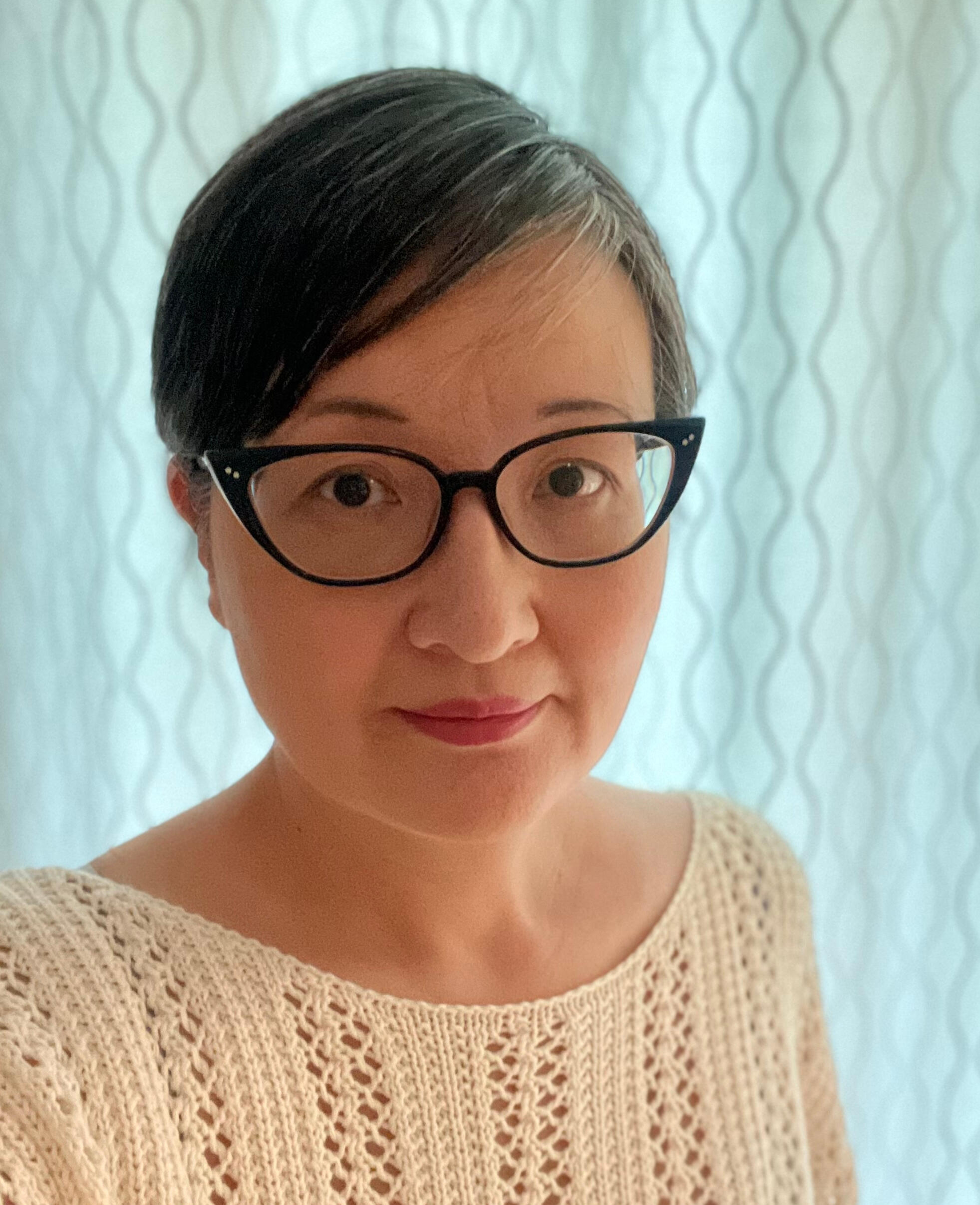 Kat Kruger An image of me, Kat Kruger, with black hair in a pixie cut. I have black-rimmed cats eye glasses and am wearing a cream knit top.