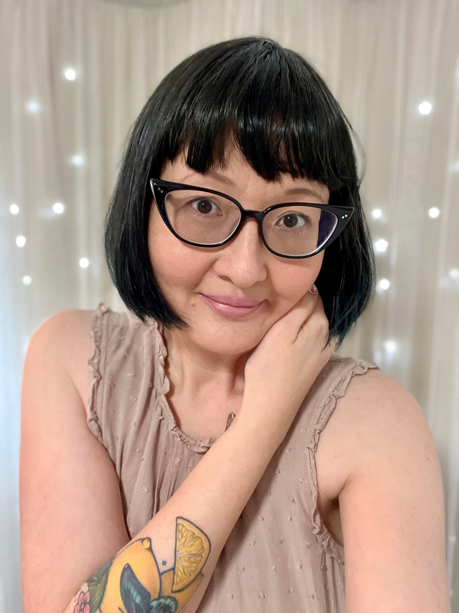 An image of me, Kat Kruger, with short teal green hair with baby bangs. I have black-rimmed cats eye glasses and am wearing a colorful moth print dress with ties at the shoulders.