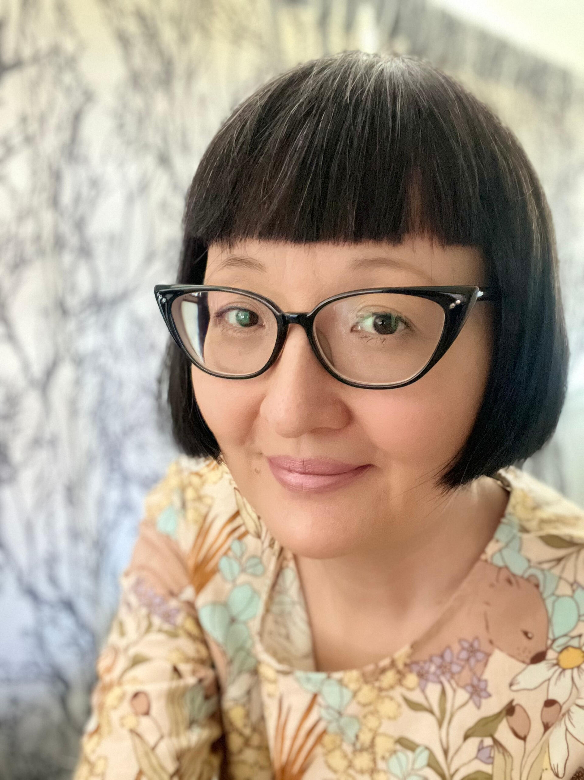 An image of me, Kat Kruger, with short black hair and bangs. I have black-rimmed cats eye glasses and am wearing a beige top with a floral and wombat print.